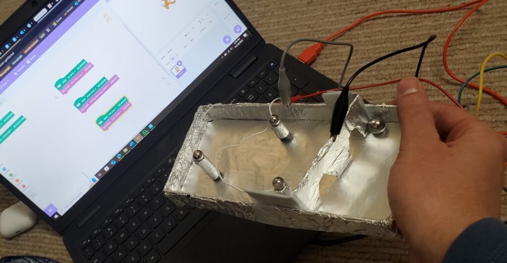 A laptop computer, connected through wires to a metallic game board held in a hand